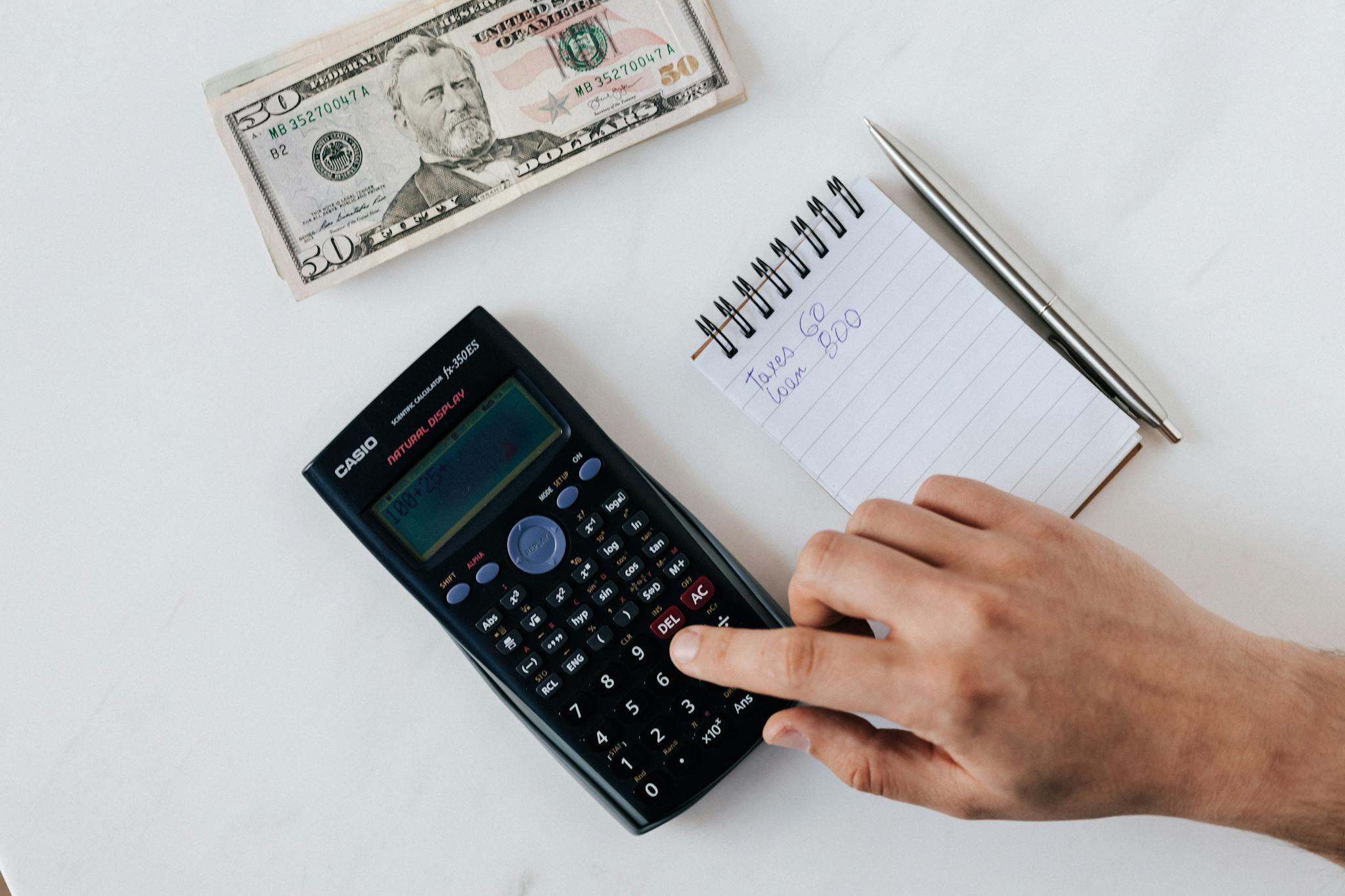 Top view of crop unrecognizable economist calculating on calculator on table with notepad and pen near pile of dollar banknotes on marble table